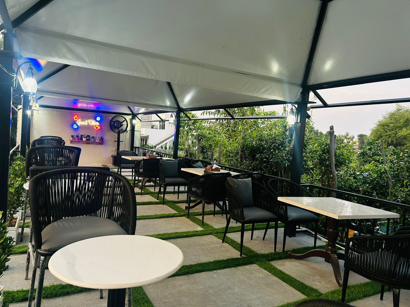 An outdoor cafe with tables and chairs, a neon sign, and a view of greenery.