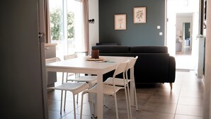 A modern living room with a white dining table, white chairs, a black sofa, and a grey accent wall with framed artwork. - Résidence des Sables (Mably)