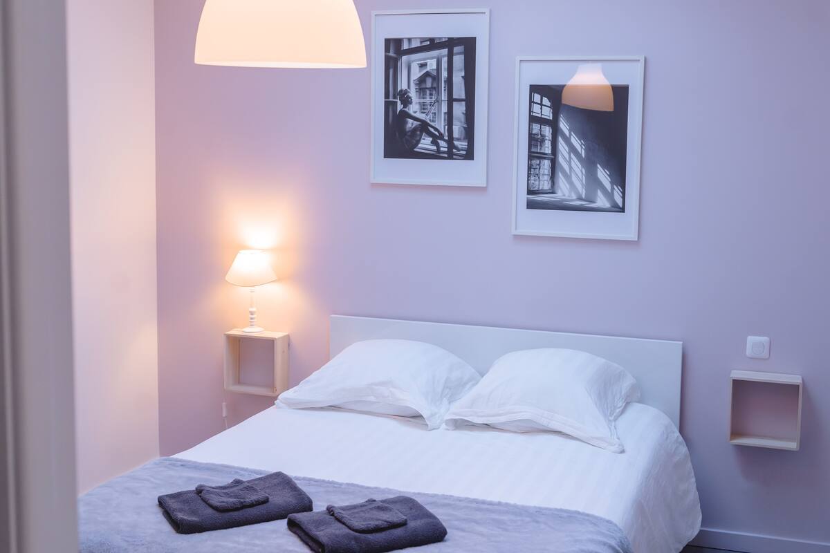 A neatly made bed with white linens and two folded towels, a bedside lamp, and two framed pictures on the wall.