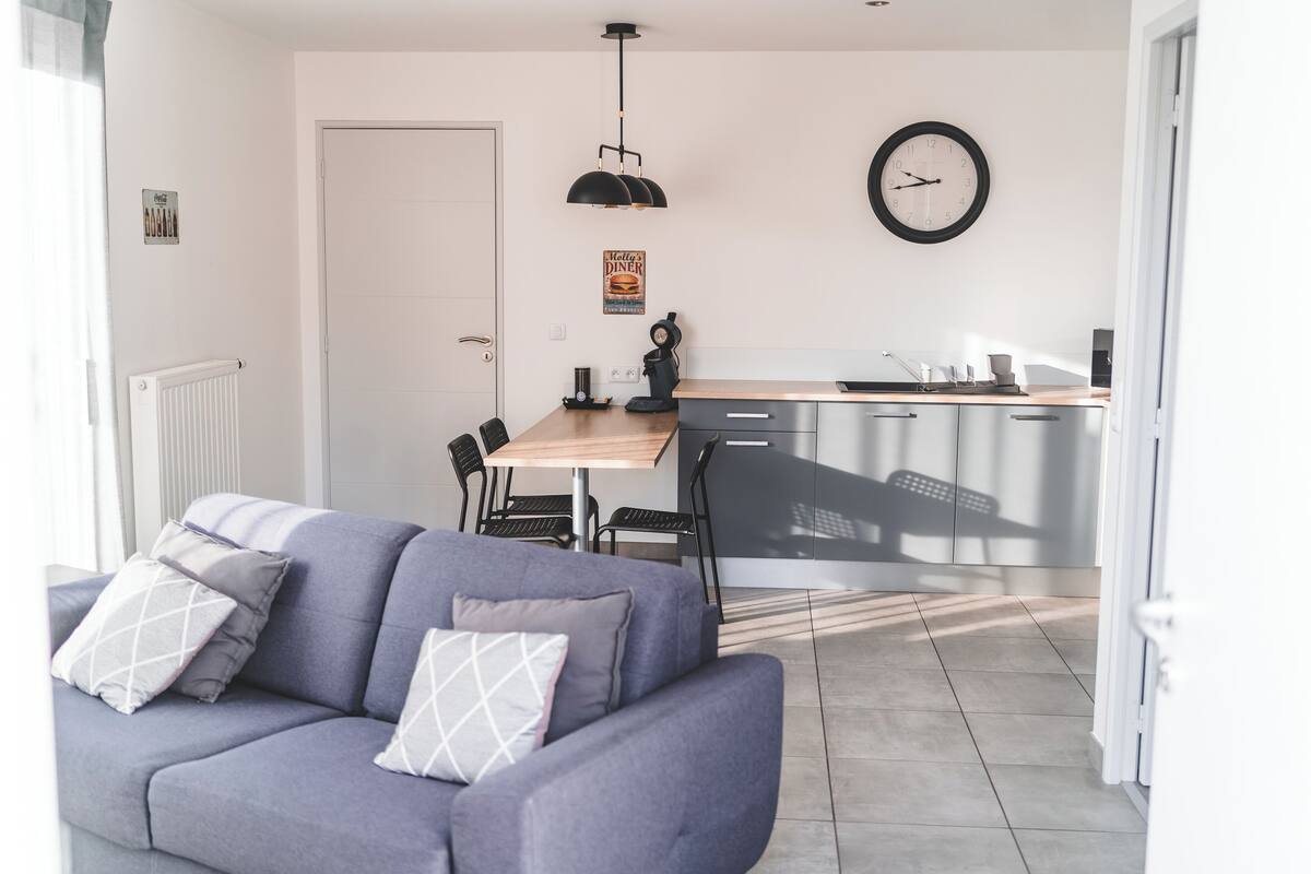 A modern living room with a grey sofa, a wooden dining table, and a kitchen area with a clock and a poster on the wall.