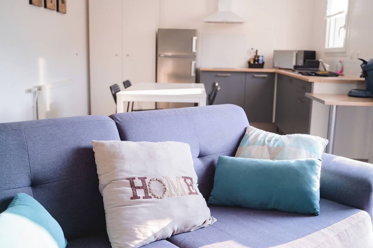 A modern living room with a sofa, a kitchen in the background, and a dining area.