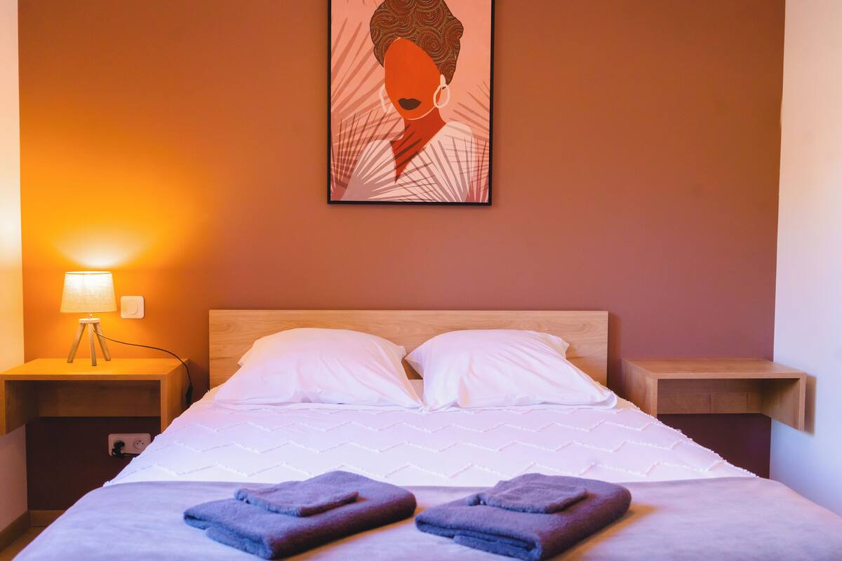 A neatly made bed with white linens and two pillows, a bedside lamp, and a wall-mounted artwork.