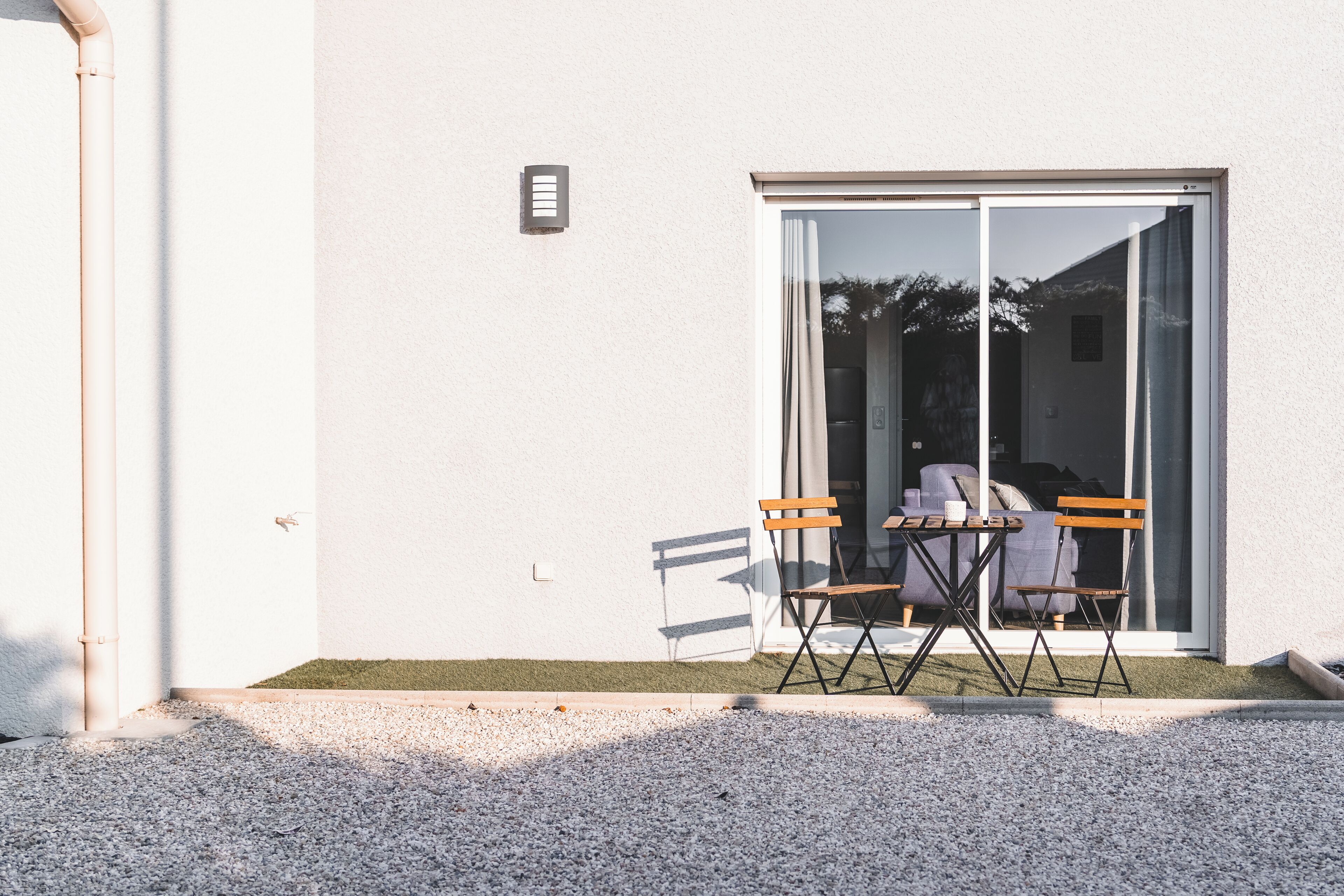 Eine Terrasse mit Tisch und zwei Stühlen, eine Glastür und eine kleine Rasenfläche.