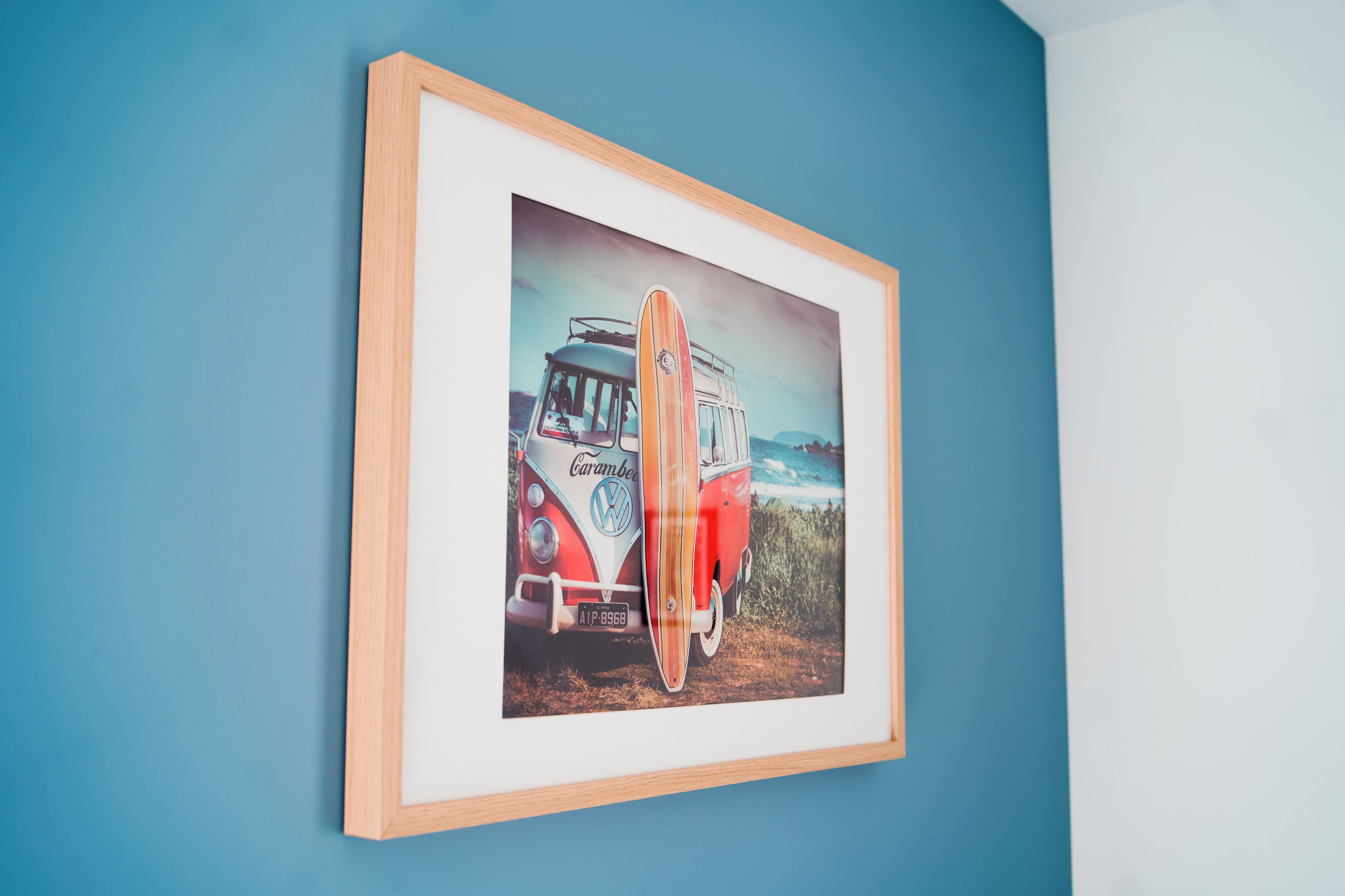 A framed picture of a vintage VW camper van with a surfboard on the back, mounted on a blue wall.