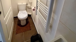 A small, modern bathroom with a toilet, a white radiator, and a black trash bin. - Comfortable 2-room apartment, independent access & PMR. (Épinay-sur-Seine)