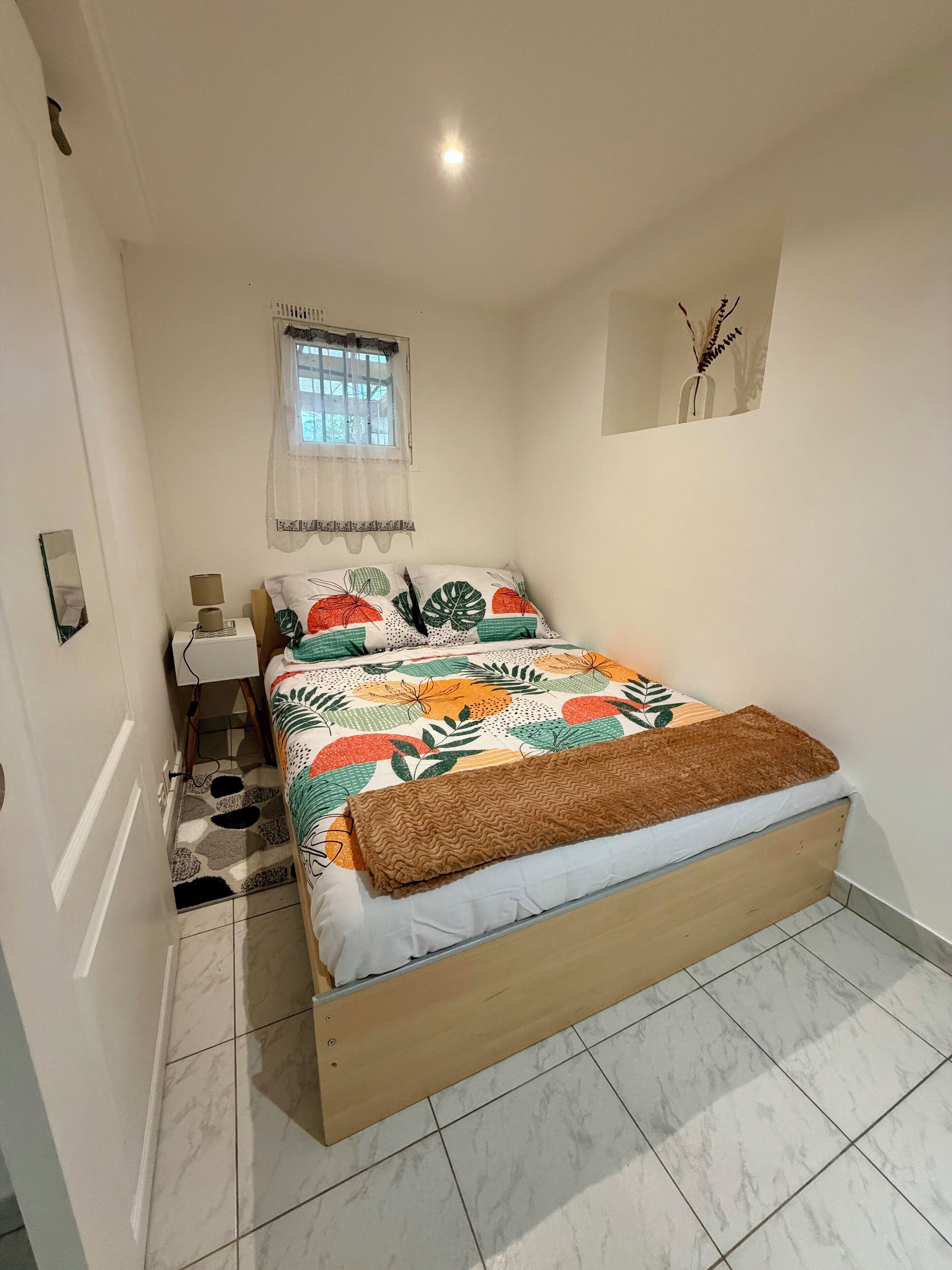 A bedroom with a bed, a nightstand, a lamp, a window with curtains, and a small wall decoration.