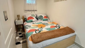A bedroom with a bed, a nightstand, a lamp, a window with curtains, and a small wall decoration. - Comfortable 2-room apartment, independent access & PMR. (Épinay-sur-Seine)