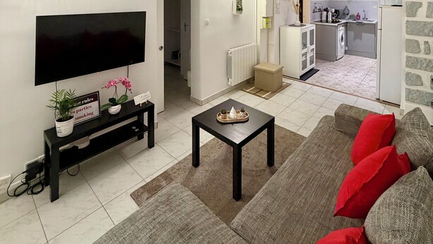 A living room with a flat-screen TV, a coffee table, and a sofa with red cushions. There is a kitchen area in the background with a washing machine and a refrigerator. - Comfortable 2-room apartment, independent access & PMR. (Épinay-sur-Seine)