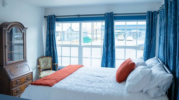 A bedroom with a large window, a wooden cabinet, a chair, and a bed with white bedding and red pillows. - Palmer Suite 101, Hyland Hotel (Palmer)