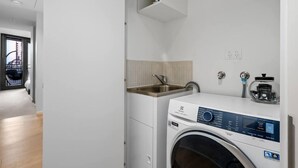 A compact laundry room with a washing machine, a sink, and a small shelf above the machine. - Stylish Arts Apt | Lift | Gym | Sauna (Southbank)