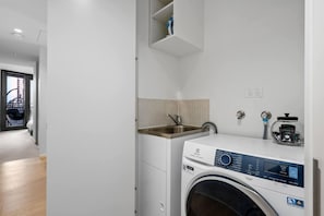 A compact laundry room with a washing machine, a sink, and a small shelf above the machine. - Stylish Arts Apt | Lift | Gym | Sauna (Southbank)