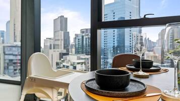 A dining area with a view of a cityscape through large windows, a round table with black bowls and a wine glass, and a white chair.