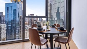 A dining area with a table set for four, large windows offering a city view, and modern chairs. - Stylish Arts Apt | Lift | Gym | Sauna (Southbank)