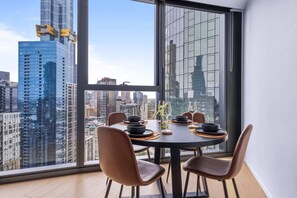 A dining area with a table set for four, large windows offering a city view, and modern chairs. - Stylish Arts Apt | Lift | Gym | Sauna (Southbank)