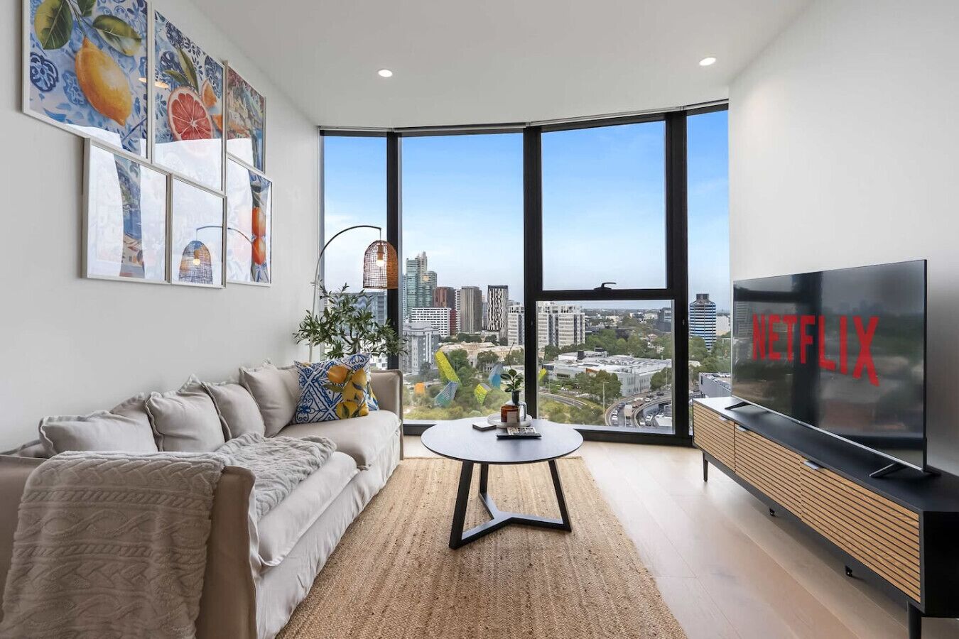 A modern living room with a large TV, a sofa, a coffee table, and a city view through large windows.