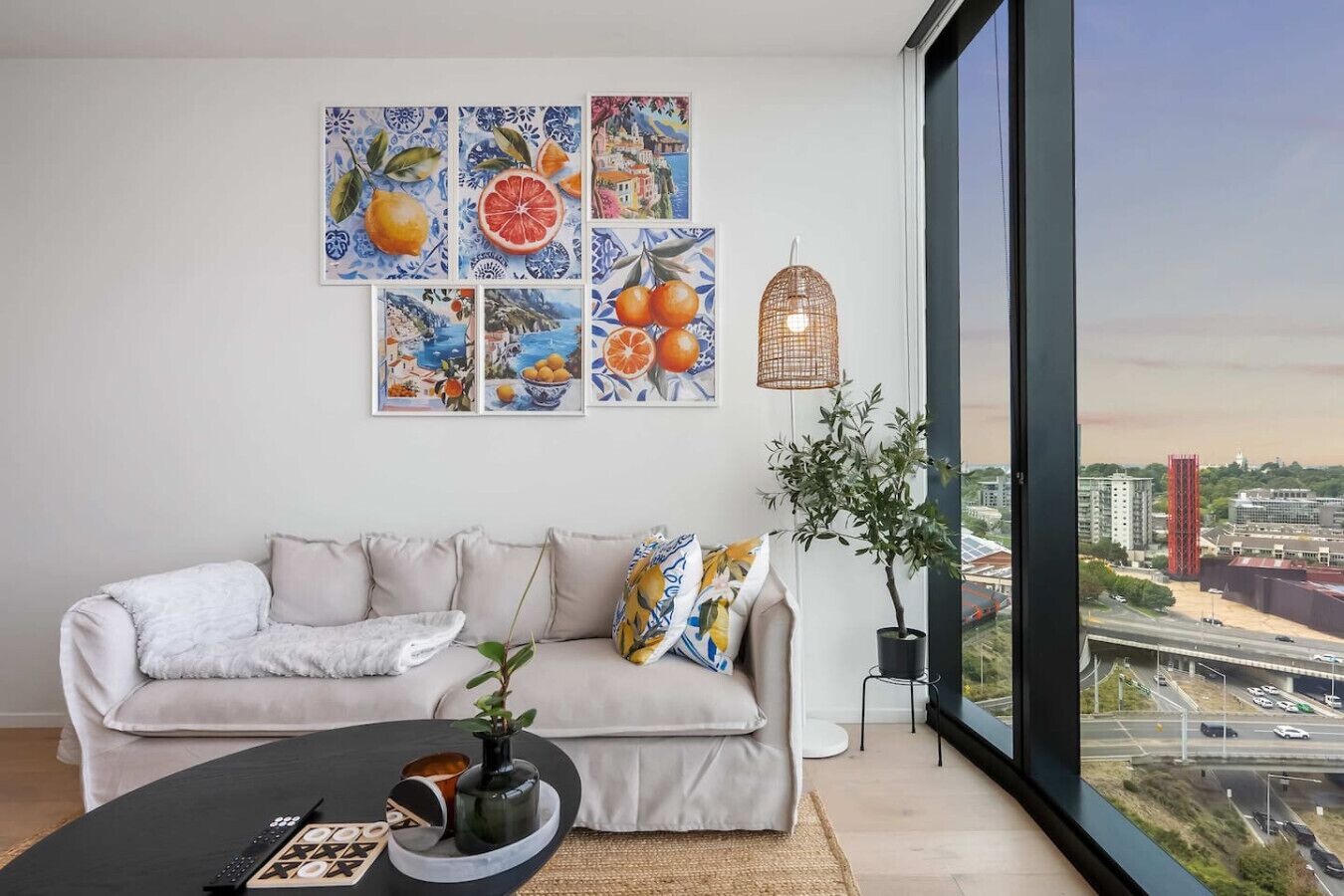 A modern living room with a sofa, artwork, a plant, and a view of a cityscape through a large window.