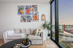 A modern living room with a sofa, artwork, a plant, and a view of a cityscape through a large window. - Amalfi Chic | Pool | Gym | Sauna (Southbank)
