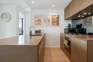 Eine moderne Küche mit Kochinsel, Edelstahlgeräten und Holzausstattung. Drei gerahmte Kunstwerke hängen an der Wand, und eine Uhr ist links im Bild zu sehen.