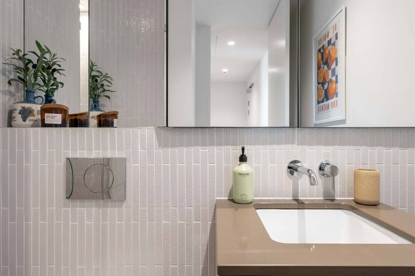 Un baño con pared de azulejos blancos, un espejo, un lavabo con grifo cromado, un dispensador de jabón y un perchero para toallas.