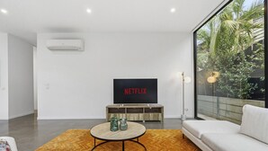 A modern living room with a flat-screen TV, a sofa, a coffee table, and a view of greenery outside.