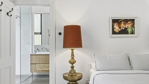 A bedroom with a bed, a bedside table with a lamp, a painting, and a bathroom visible through an open door.