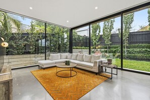 Un salon moderne avec un canapé d’angle blanc, une table basse ronde et une grande fenêtre donnant sur un jardin.