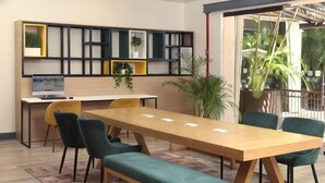 Una habitación con una larga mesa de madera, bancos acolchados verdes, sillas amarillas, un escritorio blanco con una computadora y una estantería con plantas.