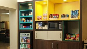 Estación autoservicio de aperitivos y bebidas con refrigerador, microondas y diferentes productos envasados.