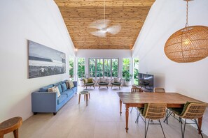 A spacious living area with a blue sofa, a wooden dining table, wicker chairs, and a television. - The Oasis Villa (Kerobokan)
