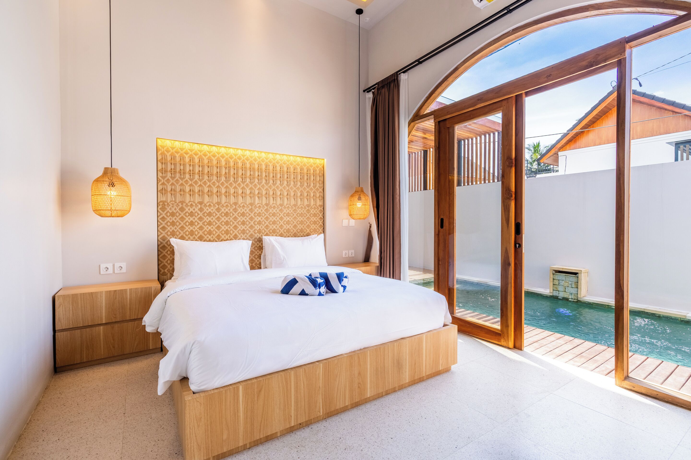 A modern bedroom with a large bed, a wooden headboard, and a view of a swimming pool through glass doors.