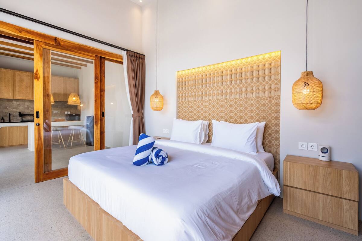 A modern hotel room with a large bed, a wooden headboard, a bedside table with a speaker, and a sliding glass door leading to a kitchen area.