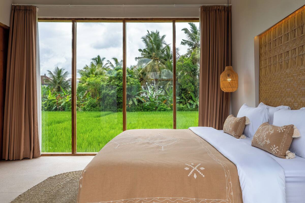 A bedroom with a large bed, a view of a green field, and a woven wall panel.