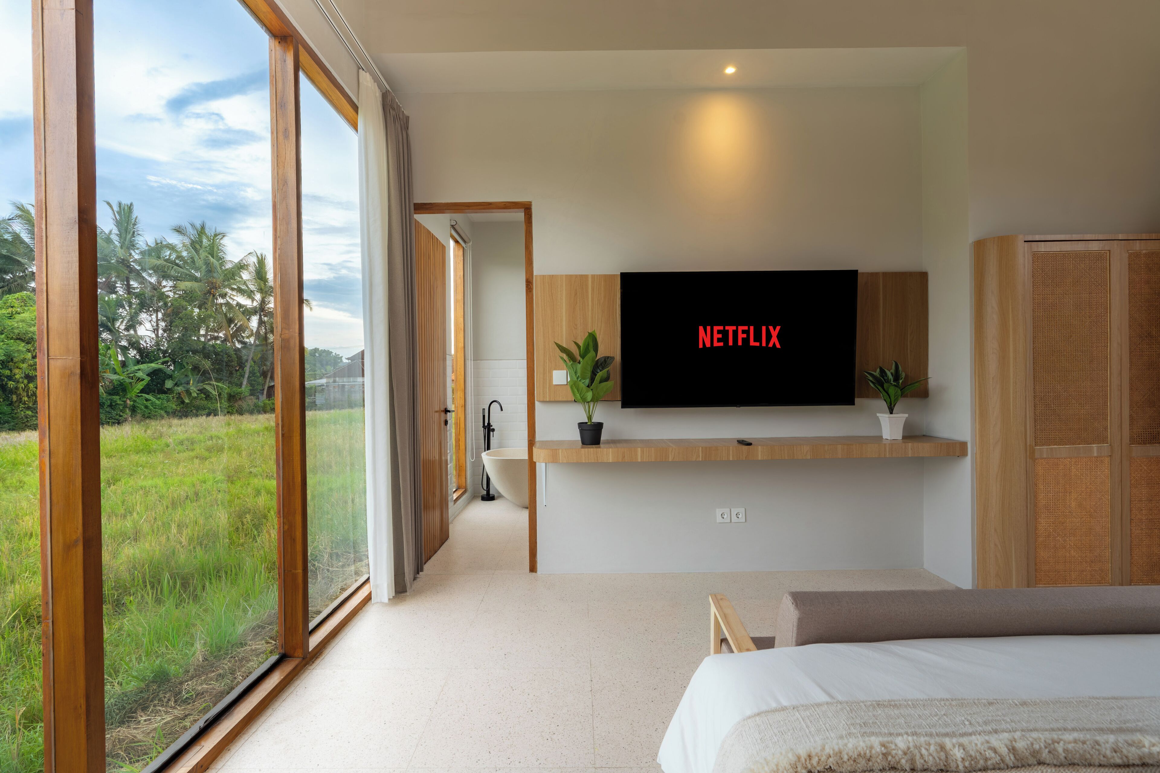 A bedroom with a large window, a bed, a TV mounted on the wall, and a small plant on the bedside table.