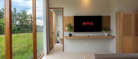 A bedroom with a large window, a bed, a TV mounted on the wall, and a small plant on the bedside table.