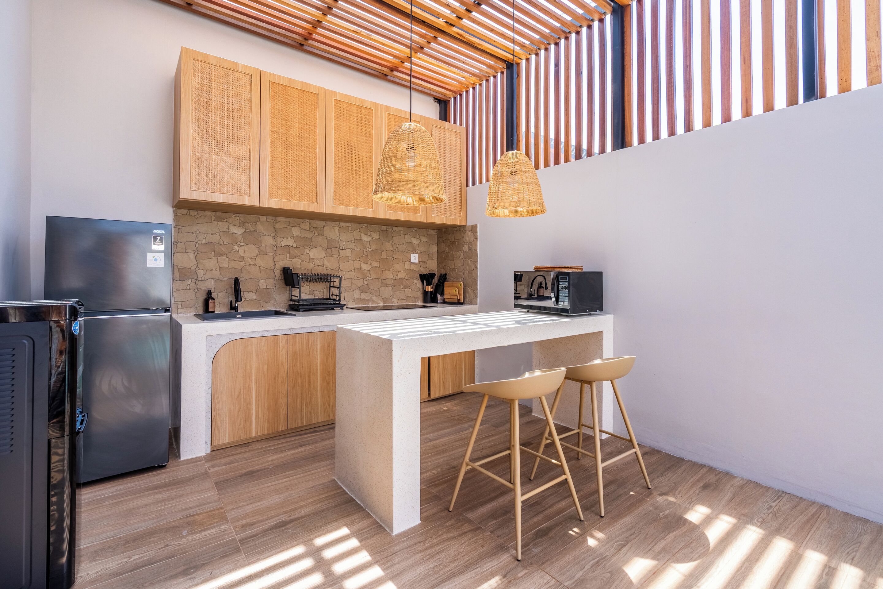 A modern kitchen with a stone backsplash, wooden cabinets, a stainless steel refrigerator, a coffee machine, and two hanging light fixtures.
