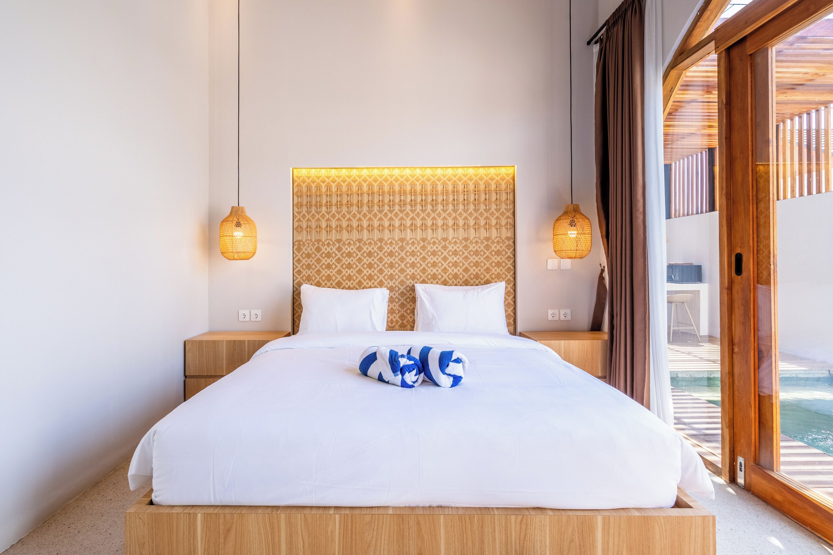A neatly made bed with white linens and a blue and white towel, a patterned headboard, and two bedside lamps.