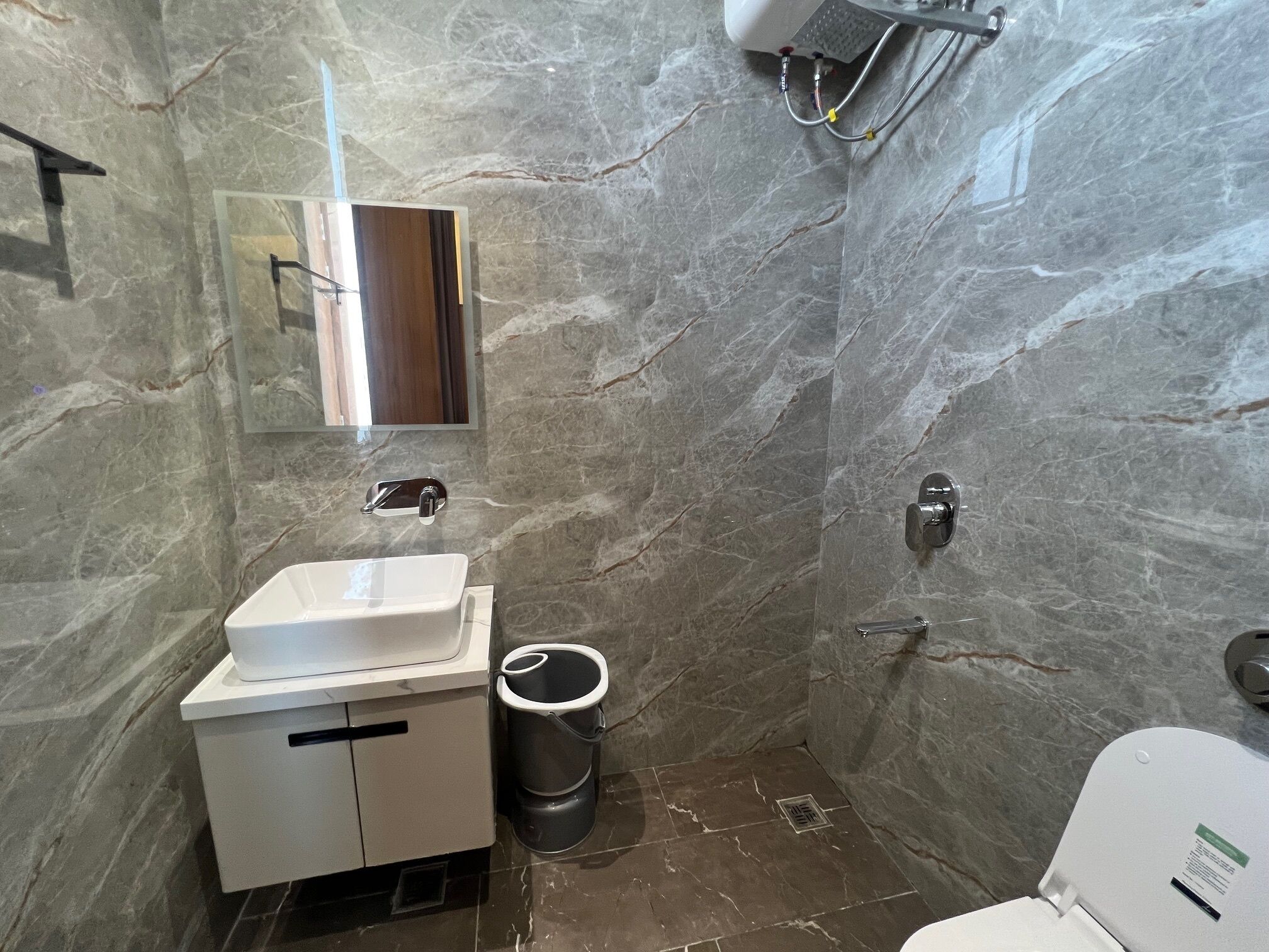 A bathroom with a marble wall, a sink, a mirror, a toilet, and a trash bin.