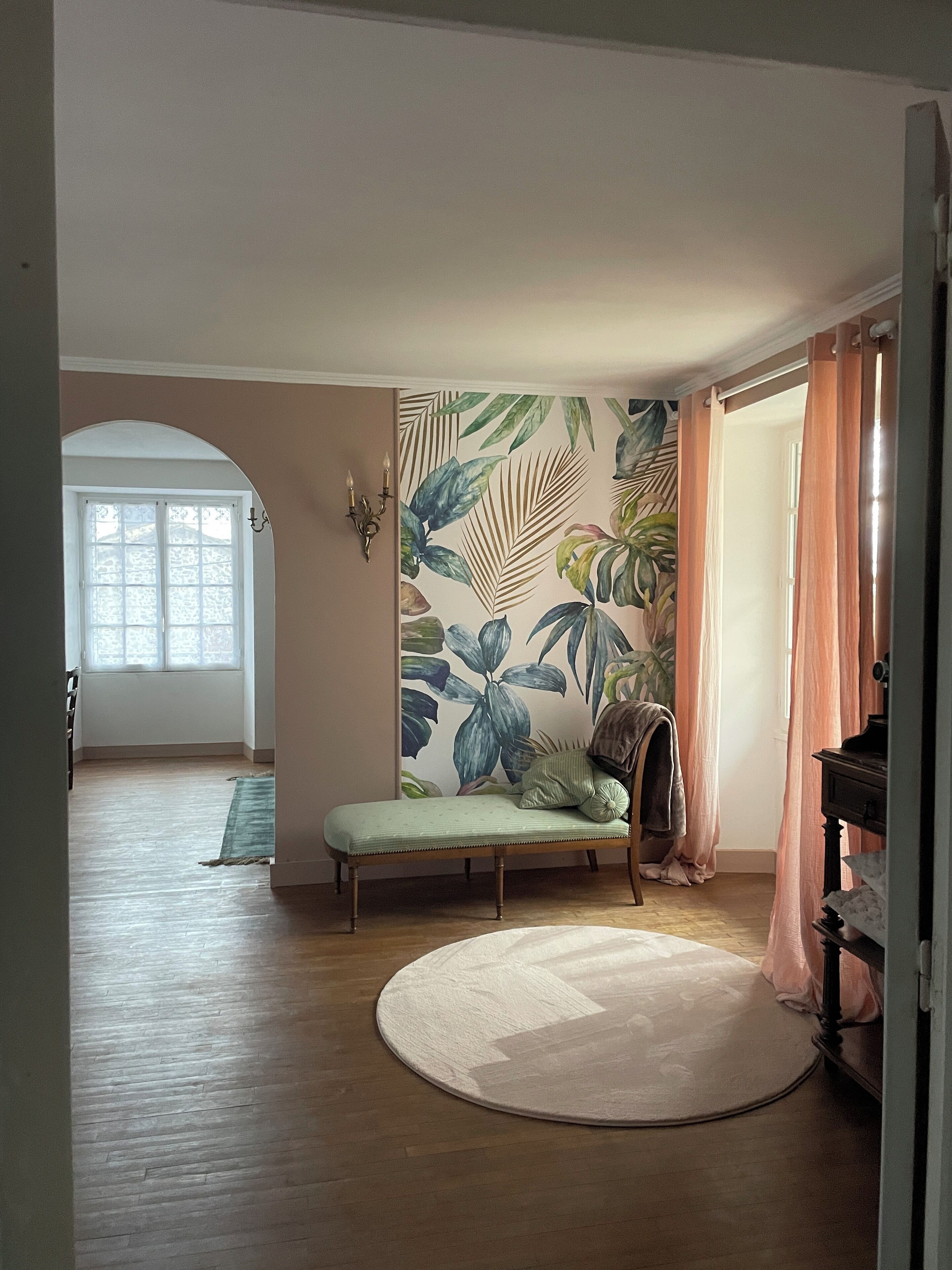 Une pièce dotée d’une grande fenêtre, d’un banc, d’un tapis rond et d’un mur orné d’un motif de feuilles tropicales.