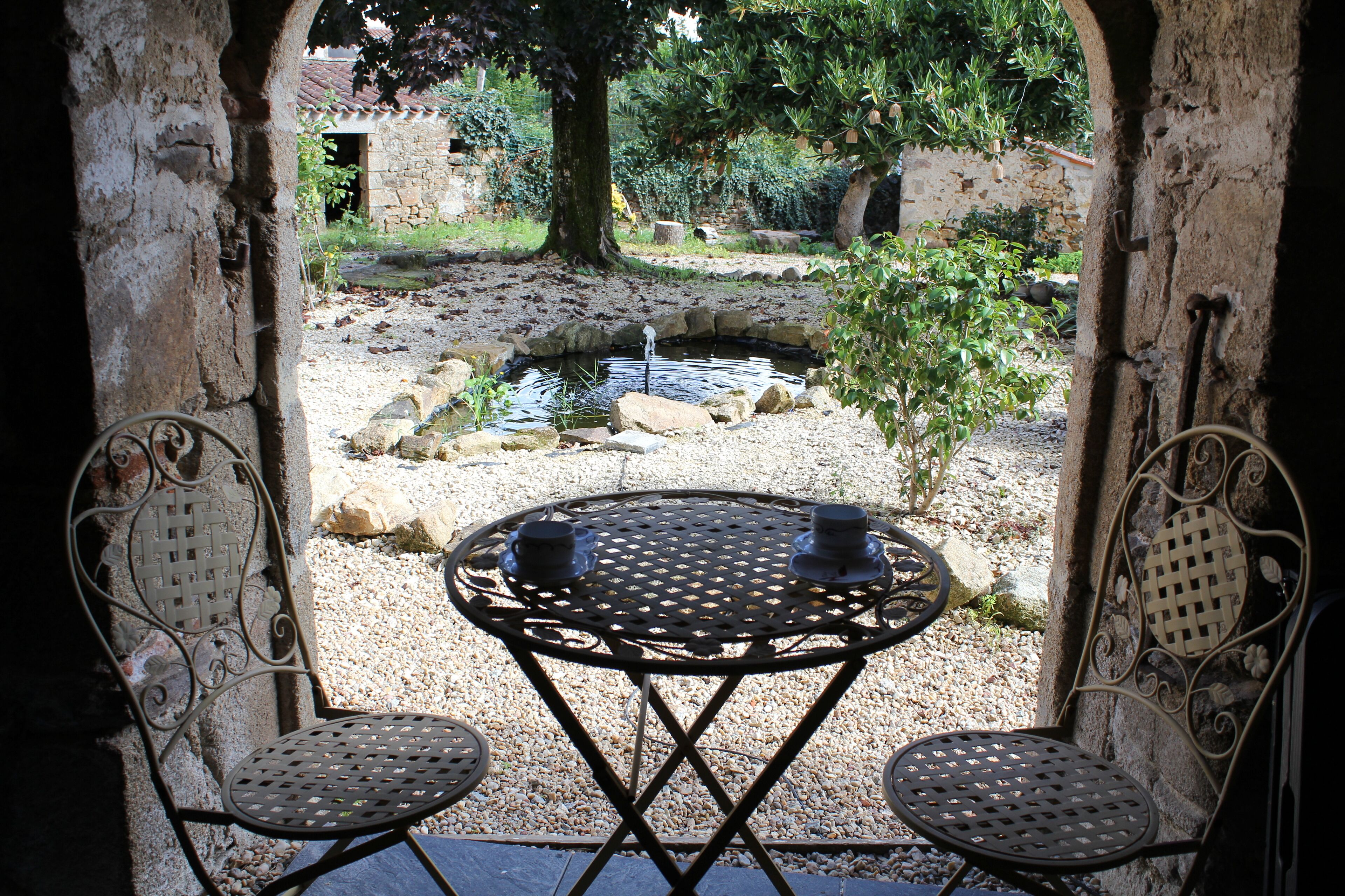 Un espace extérieur aménagé dans un style rustique, avec une table ronde en métal et deux chaises, donnant sur un petit étang et un jardin.