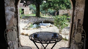 Un espace extérieur aménagé dans un style rustique, avec une table ronde en métal et deux chaises, donnant sur un petit étang et un jardin.