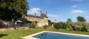 Une grande maison avec une piscine, entourée de verdure, et une petite église en arrière-plan.
