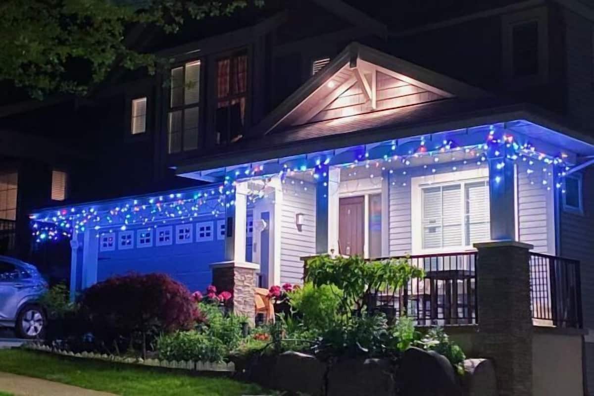 A house with a porch decorated with string lights, a car parked in the driveway, and a well-maintained garden with various plants and flowers.