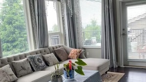 Ein Wohnzimmer mit einem beigen Sofa, gemusterten Kissen, einem Couchtisch mit blau kariertem Tischläufer und zwei Blumentöpfen.