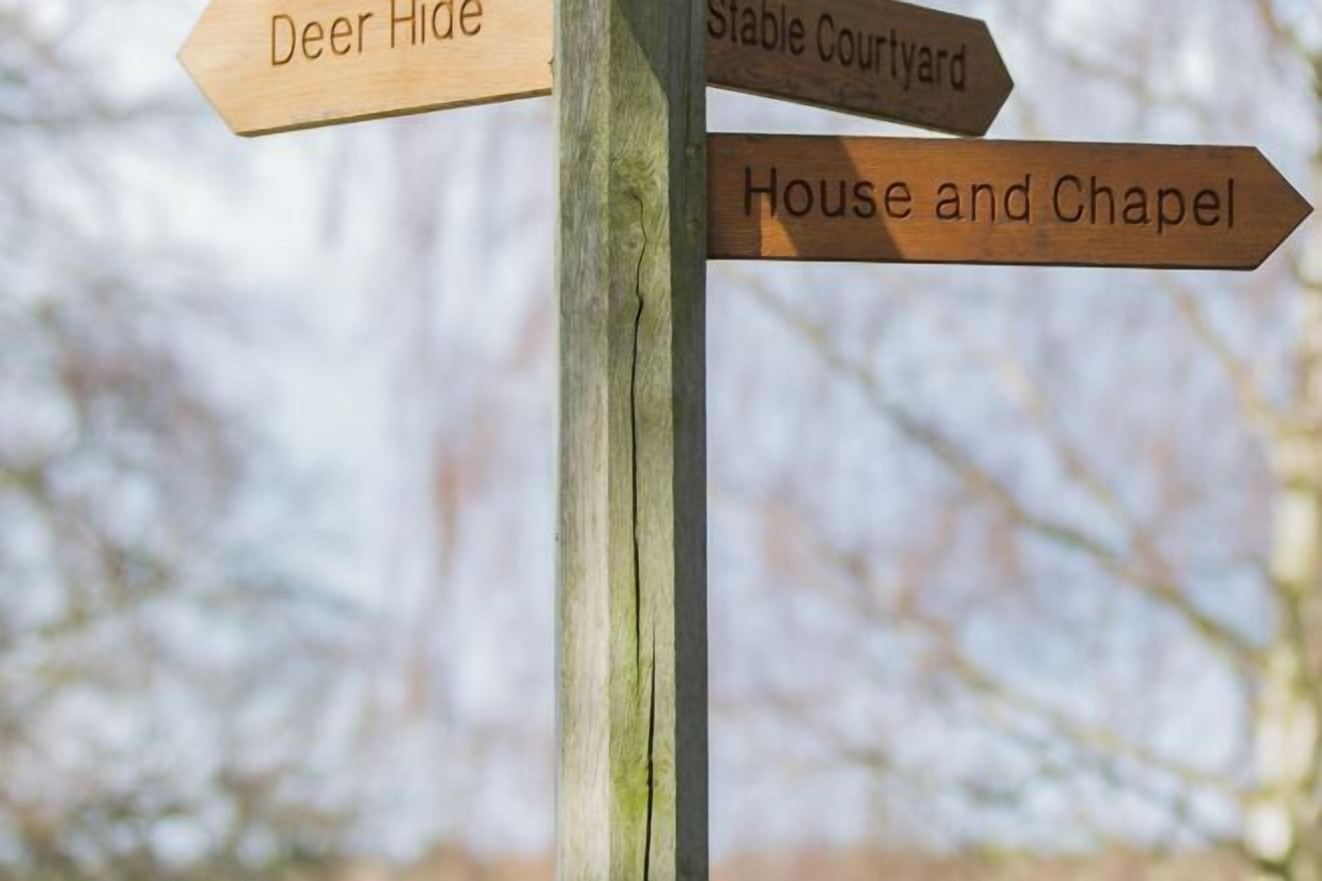 A wooden directional sign with arrows pointing to 'Deer Hide', 'Stable Courtyard', and 'House and Chapel'.