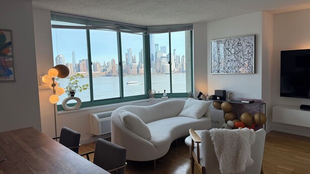 A modern living room with a white sofa, a dining table, and chairs. A large window offers a view of a city skyline and a river. - Newport Apartment (Jersey City)