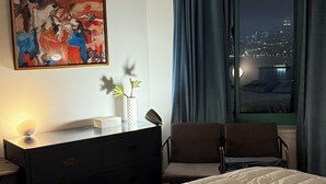 A hotel room with a bed, a desk with a lamp, a painting on the wall, and a view of the city at night. - Newport Apartment (Jersey City)