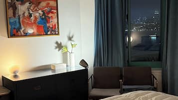 Habitación de hotel con cama, un escritorio con lámpara, un cuadro en la pared y vista a la ciudad de noche.