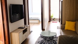 Ein modernes Wohnzimmer mit einem Flachbildfernseher, einem Sofa mit gelbem Kissen, einem Glastisch und Blick auf ein Schlafzimmer mit Bett und Fenster.