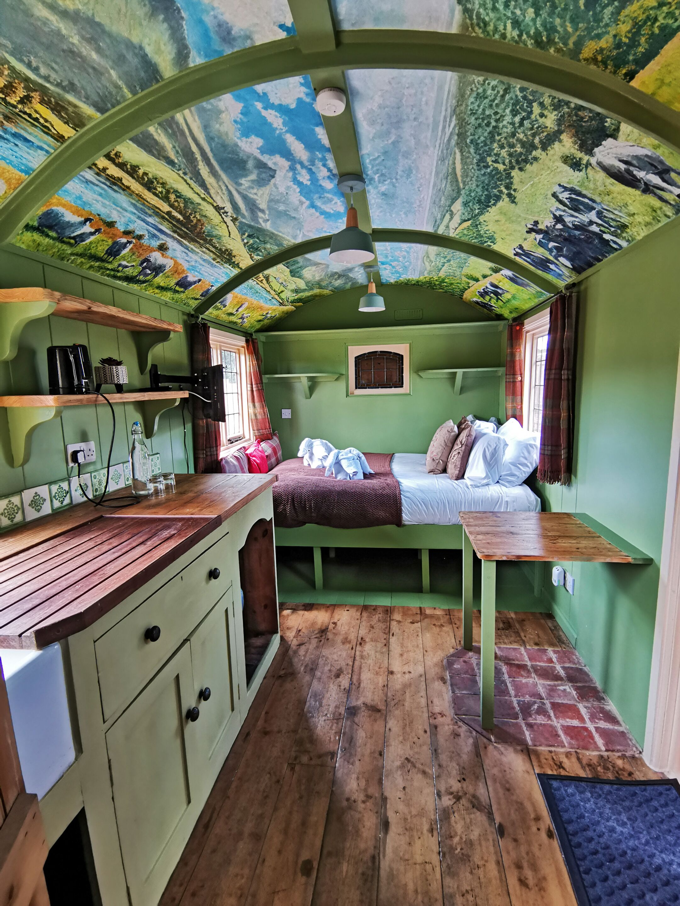 Una habitación acogedora con un mural de un paisaje, una cama, una mesita y un ventanal con cortinas.