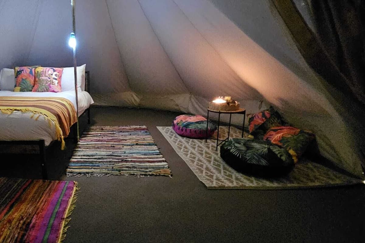 Een knus ingerichte tent met een bed, een bedlamp, een kamerkleed met patroon en een kleine tafel met een kaars.